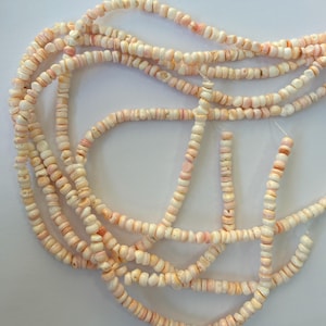 May include: A long strand of small, irregularly shaped, pale pink and white shell beads. The beads are strung together on a thin, clear filament, creating a continuous, flexible line of natural, textured, and organic-looking circular components.