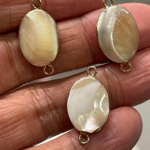 May include: Three oval-shaped shell charms with gold jump rings. The shells are iridescent with white, cream, and light brown tones.
