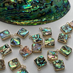 May include: A collection of iridescent abalone shell pendants with gold-toned jump rings. The pendants are square shaped and have a variety of colors, including green, blue, and purple.