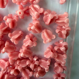 May include: A collection of small, light pink, flower-shaped beads. Each bead has a detailed, multi-petal design with a glossy finish. The beads are scattered, suggesting they are for crafting or jewelry making.