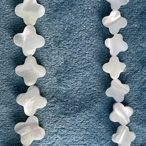 May include: Two strands of white, iridescent, shell beads in the shape of a four-leaf clover. The beads are strung on a clear cord and are displayed against a blue background.