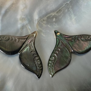 May include: Two iridescent, dark gray whale tail pendants. Each pendant features a detailed, carved floral design. The pendants are made of a material that reflects light, creating a shimmering effect. The overall theme is nautical and decorative.