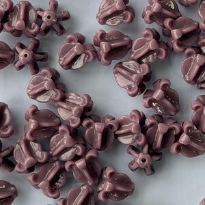 May include: Close-up of numerous mauve-colored beads. The beads have a unique, flower-like design with multiple petals and a central hole. The beads are scattered across a light blue surface, creating a visually appealing pattern. Ideal for jewelry making.