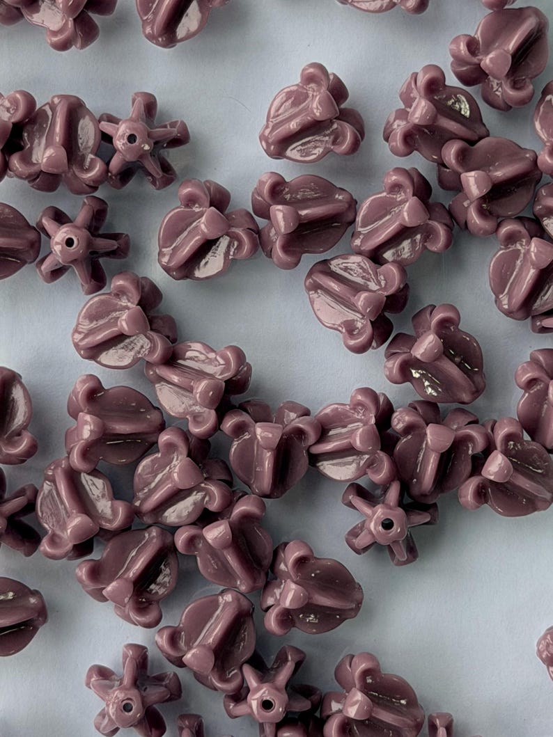 May include: Close-up of numerous mauve-colored beads. The beads have a unique, flower-like design with multiple petals and a central hole. The beads are scattered across a light blue surface, creating a visually appealing pattern. Ideal for jewelry making.