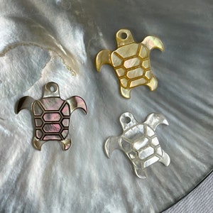 May include: Three turtle shaped charms made of mother of pearl. The charms are in different colors: gold, pink, and white.