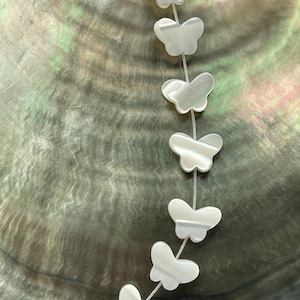May include: A strand of white mother of pearl butterfly beads. The beads are strung on a white thread and are arranged in a vertical line.