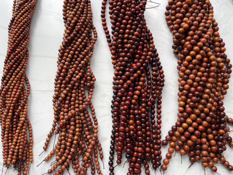 May include: Four strands of round, polished brown wooden beads arranged vertically. Each strand consists of numerous small, uniform spheres strung together on a thin black cord, showcasing varying shades of natural wood grain and texture.
