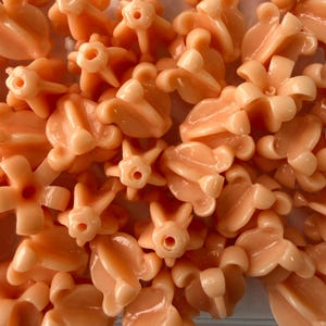 May include: A collection of small, peach-colored plastic flower-shaped beads. The beads have a glossy finish and various designs, including four-petal and six-petal flower shapes. The beads are scattered, filling the frame.