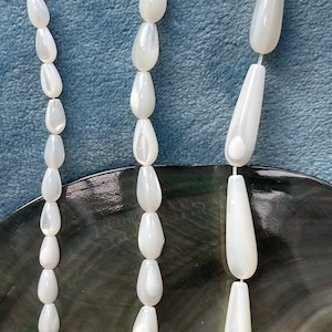 May include: A strand of white pearlescent teardrop-shaped beads. The beads are strung on a white cord and are arranged in a vertical line. The beads are iridescent and have a smooth, polished finish.