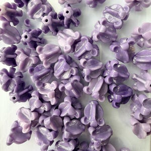 May include: A collection of light purple flower-shaped beads. Each bead has a detailed, textured design with multiple petals and a central hole. The beads are scattered across a white surface, creating a visually appealing arrangement.