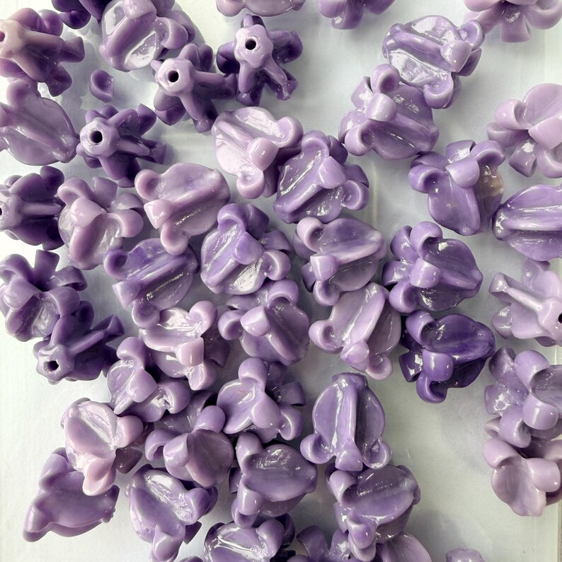 May include: A collection of light purple flower-shaped beads. Each bead has a detailed, textured design with multiple petals and a central hole. The beads are scattered across a white surface, creating a visually appealing arrangement.