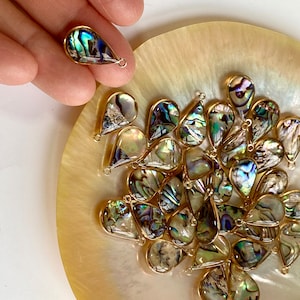 Könnte beinhalten: Eine Sammlung von regenbogenfarbenen Abalone-Muschel-Tropfenanhängern mit goldfarbenen Sprungringen. Die Anhänger liegen auf einer Perlmuttschale.