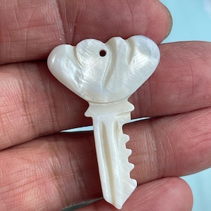 May include: A pearlescent white key pendant with a flower-shaped top and a key-shaped bottom. The key has a small hole at the top for hanging. The pendant is made of a shiny, iridescent material.