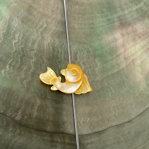 May include: A decorative pin with a small, iridescent fish design in yellow and cream hues. The fish is attached to a long, silver-coloured pin. The background is a mottled, green-grey surface.