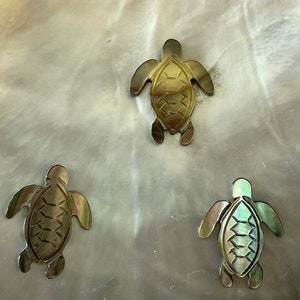 May include: Three iridescent green and gold turtle shaped ornaments on a white background.