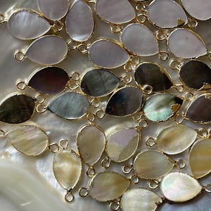 May include: A collection of pearlescent and iridescent shell teardrop-shaped pendants with gold-toned jump rings. The pendants are arranged on a white, textured surface.