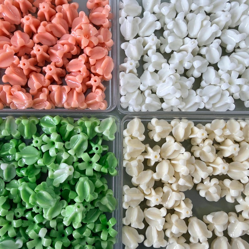 May include: Overhead shot of four compartments filled with decorative beads. The beads are in shades of peach, white, green, and cream. Each bead has a unique floral design, perfect for jewelry making or craft projects.