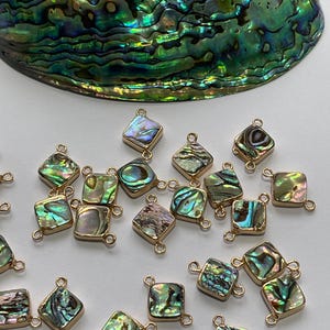 Puede incluir: Cuadrados de concha de abulón con bordes y bucles de metal dorado. Los cuadrados iridiscentes muestran tonos de verde, azul y morado. La imagen también muestra una gran concha de abulón en el fondo. Adecuado para la fabricación de joyas.