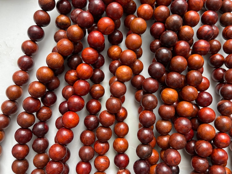May include: A collection of round, polished wooden beads in various shades of deep reddish-brown and dark brown. The beads are arranged in multiple vertical strands, showcasing natural wood grain patterns and a smooth, uniform spherical shape.