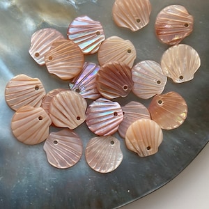 Peut inclure: Un ensemble de boutons en forme de coquillage rose et irisé avec un trou au centre. Les boutons sont disposés sur une surface gris foncé.