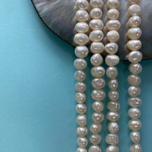 Op de afbeelding: Een streng witte barokparels, met een licht onregelmatige vorm, gerangschikt in een rij op een parelmoer oppervlak.