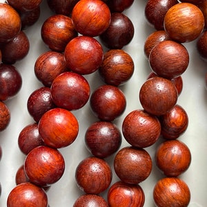 May include: A collection of polished, spherical wooden beads arranged in vertical strands. Each bead displays a rich, deep reddish-brown color with a visible natural wood grain pattern, smooth texture, and a glossy finish throughout the surface.