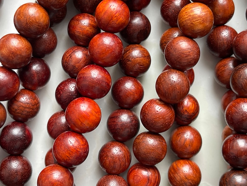 May include: A collection of polished, spherical wooden beads arranged in vertical strands. Each bead displays a rich, deep reddish-brown color with a visible natural wood grain pattern, smooth texture, and a glossy finish throughout the surface.