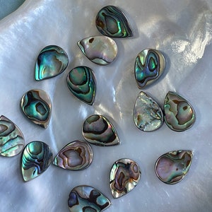 May include: A selection of teardrop-shaped abalone shell cabochons, displaying iridescent hues of green, blue, and purple. The cabochons are presented on a pearlescent, textured surface, highlighting their natural beauty and unique patterns. Perfect for jewellery making.