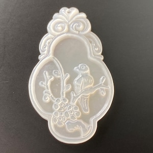 Op de afbeelding: Een witte, sierlijke, ovale hanger met een vogel die op een tak met bloemen zit. De hanger heeft een decoratieve bovenkant met een lus om op te hangen.