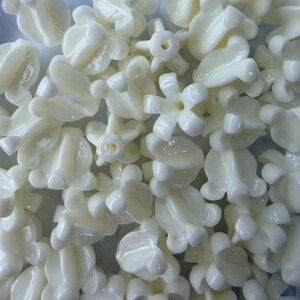 May include: A collection of off-white, flower-shaped beads. The beads have a glossy finish and various floral designs, including five-petal and tulip-like shapes. These beads are likely used for jewelry making or craft projects.