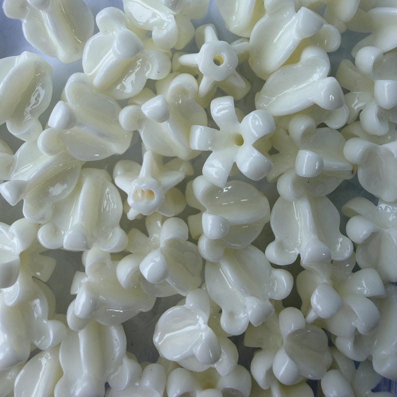 May include: A collection of off-white, flower-shaped beads. The beads have a glossy finish and various floral designs, including five-petal and tulip-like shapes. These beads are likely used for jewelry making or craft projects.