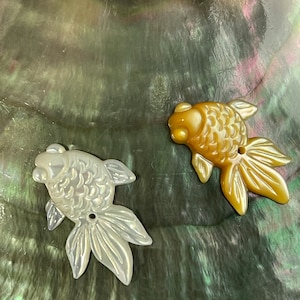 Może przedstawiać: Dwa rzeźbione wisiorki w kształcie ryb wykonane z perłowej muszli. Jedna ryba jest biała, a druga złota.