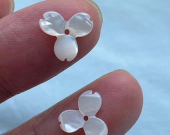 Könnte beinhalten: Zwei kleine, irisierende, weiße blütenförmige Perlen. Jede Perle hat drei Blütenblätter und ein kleines Loch in der Mitte. Die Perlen werden zwischen den Fingern gehalten, was ihr zartes Design und ihren perlmuttartigen Glanz zeigt. Ideal für die Schmuckherstellung.