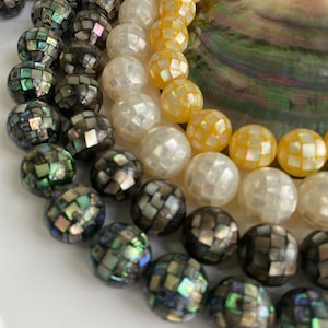 May include: A close-up of three strands of round, iridescent beads. The beads are made of mother-of-pearl and have a mosaic pattern. The beads are in shades of black, white, and yellow.