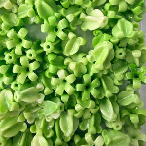 May include: A collection of light green, glossy, flower-shaped beads and leaf-shaped beads. The beads have a smooth, reflective surface and are scattered across the image. The floral beads have a five-petal design with a hole in the center.