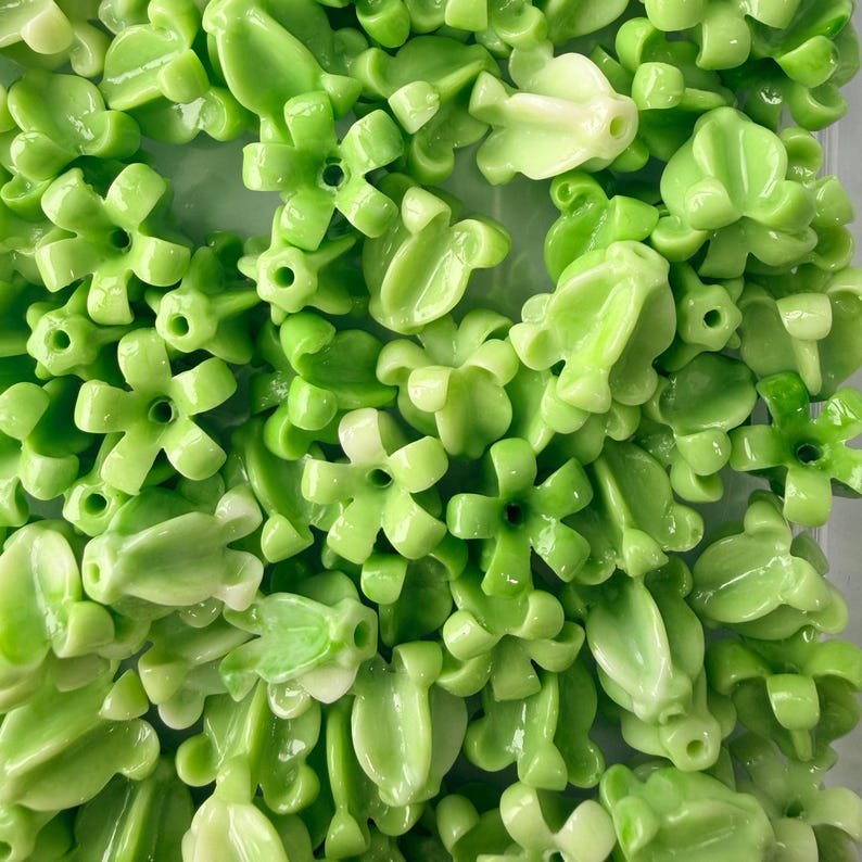 May include: A collection of light green, glossy, flower-shaped beads and leaf-shaped beads. The beads have a smooth, reflective surface and are scattered across the image. The floral beads have a five-petal design with a hole in the center.