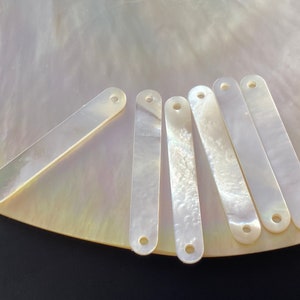 Op de afbeelding: Zes rechthoekige stukken van wit parelmoer met elk twee gaten. De stukken zijn in een waaier vorm op een witte achtergrond gerangschikt.