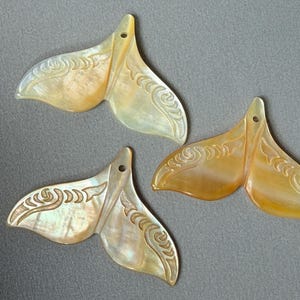 Può includere: Tre ciondoli a forma di coda di balena realizzati in madreperla iridescente. Ogni pezzo presenta una superficie liscia e lucida con motivi intagliati a spirale lungo i bordi superiori e un piccolo foro circolare al centro per l'aggancio.