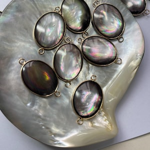 May include: A collection of eight oval-shaped iridescent mother of pearl shell charms with gold jump rings. The charms are arranged on a white mother of pearl shell surface.