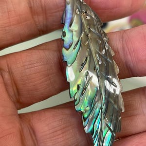 Peut inclure: Pendentif en forme de plume en coquillage d'ormeau irisé. Le pendentif présente des reflets naturels verts, bleus et argentés. La plume est finement sculptée et percée d'un trou pour l'enfilage, idéal pour un collier.