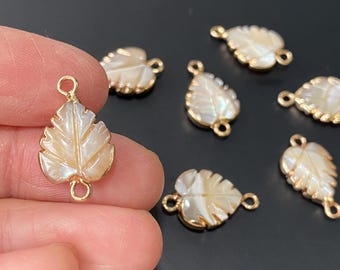 May include: Gold-coloured leaf-shaped charms with a pearlescent, off-white centre. The charms have a detailed leaf vein design and small loops for attaching to jewellery. Several charms are visible.