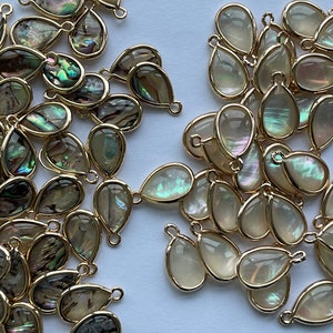 May include: A large group of iridescent teardrop-shaped pendants with gold-toned metal loops. The pendants have a pearlescent finish and are perfect for jewellery making.