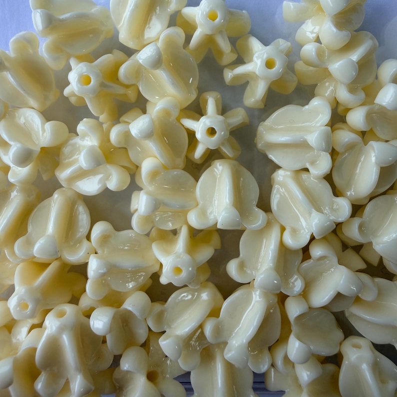 May include: A collection of ivory-colored, flower-shaped beads. The beads have a glossy finish and a variety of floral designs, including five-petal and bell-shaped forms. Each bead has a small hole for stringing.