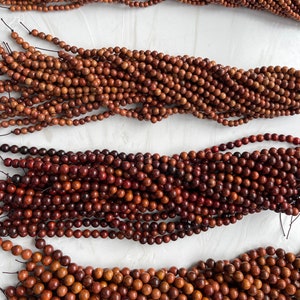 May include: Four long strands of small, round, brown wooden beads arranged horizontally. Each strand consists of numerous polished, spherical beads strung together on a thin, dark cord, showcasing natural wood grain patterns and varying brown tones.
