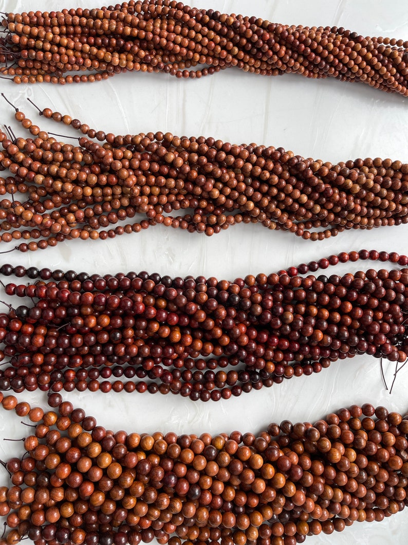 May include: Four long strands of small, round, brown wooden beads arranged horizontally. Each strand consists of numerous polished, spherical beads strung together on a thin, dark cord, showcasing natural wood grain patterns and varying brown tones.