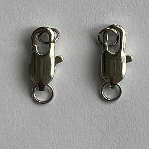 Könnte beinhalten: Zwei silberfarbene Karabinerhaken mit einem runden Ring an jeder Seite. Die Karabinerhaken sind zum Befestigen von Schmuck oder anderen Gegenständen gedacht.