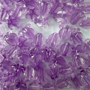 May include: A collection of translucent, light purple flower-shaped beads. The beads have a delicate, floral design with multiple petals and a clear, glossy finish. These beads are ideal for jewelry making and craft projects.