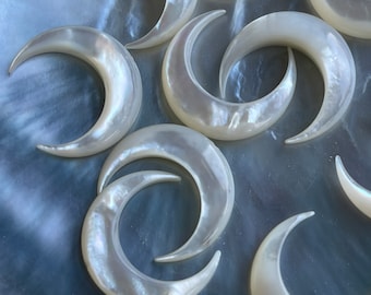 May include: A group of white crescent moon shaped charms made of mother of pearl. The charms are arranged in a random pattern on a blue background.