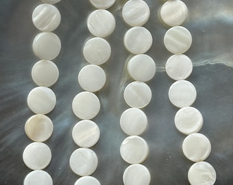 Peut inclure: Un brin de perles rondes en coquillage blanc, disposées en rangées sur une surface gris foncé. Les perles sont lisses et ont un léger éclat iridescent.