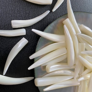 May include: A collection of white iridescent shell beads in the shape of a curved tooth. The beads are arranged on a black surface and a mother of pearl dish.